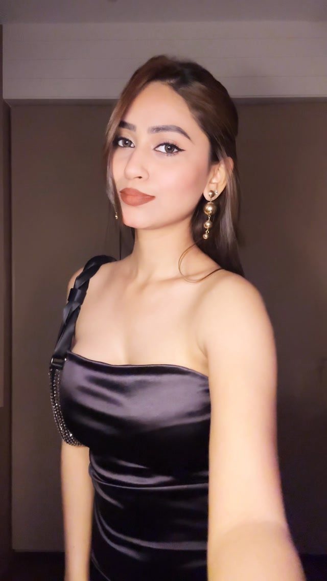 26 Classy Date Outfits From Casual to Elegant 1 Woman wearing a black satin one-shoulder dress and gold earrings posing indoors.