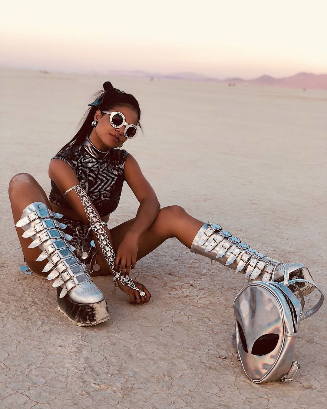 Coachella Outfits for Women: 14 Festival Looks That Work 11 Woman in futuristic silver boots and sunglasses sitting in a vast desert landscape at sunset.