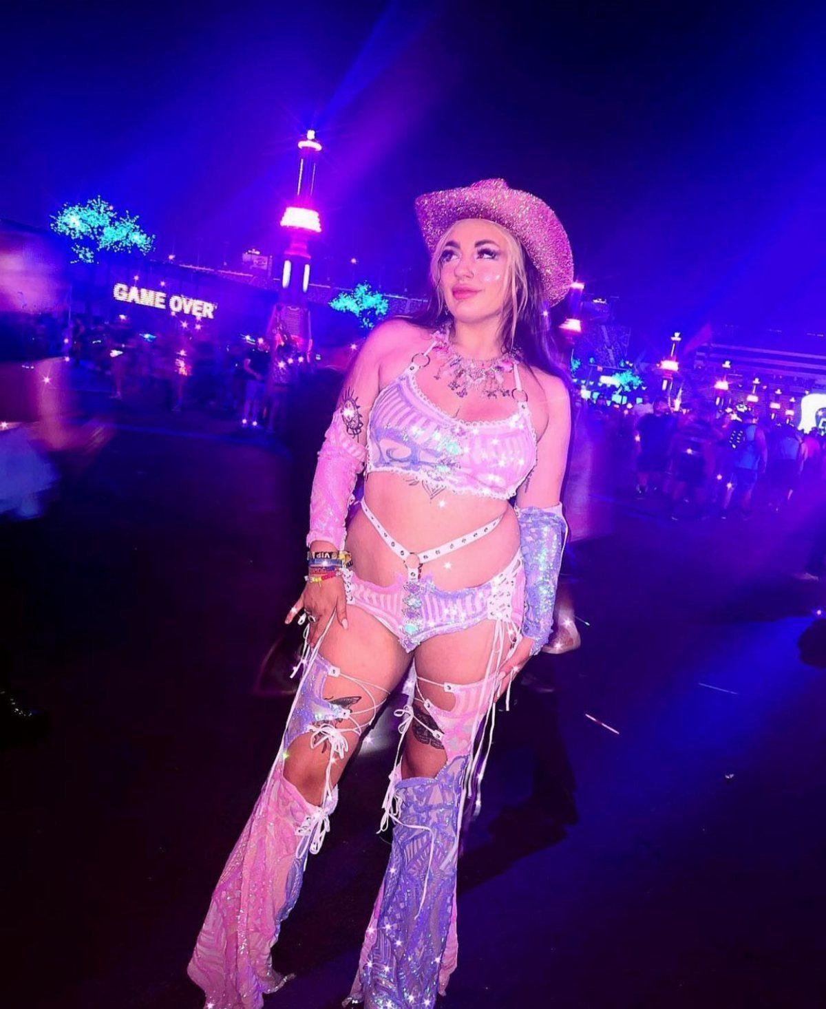 Coachella Outfits for Women: 14 Festival Looks That Work 7 Woman in sparkly pink festival outfit with chaps and cowboy hat under blue lights.