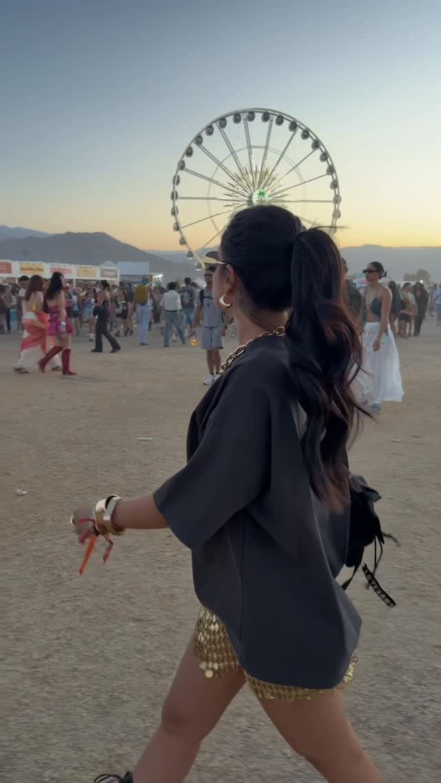 Coachella Outfits for Women: 14 Festival Looks That Work 12 Woman in gold sequined shorts and dark shirt walking through a music festival with a Ferris wheel.