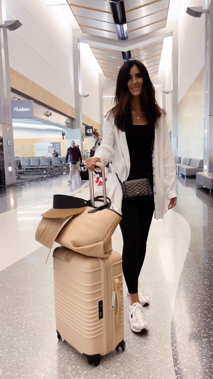 Woman in black leggings and white cardigan standing with beige suitcase in airport