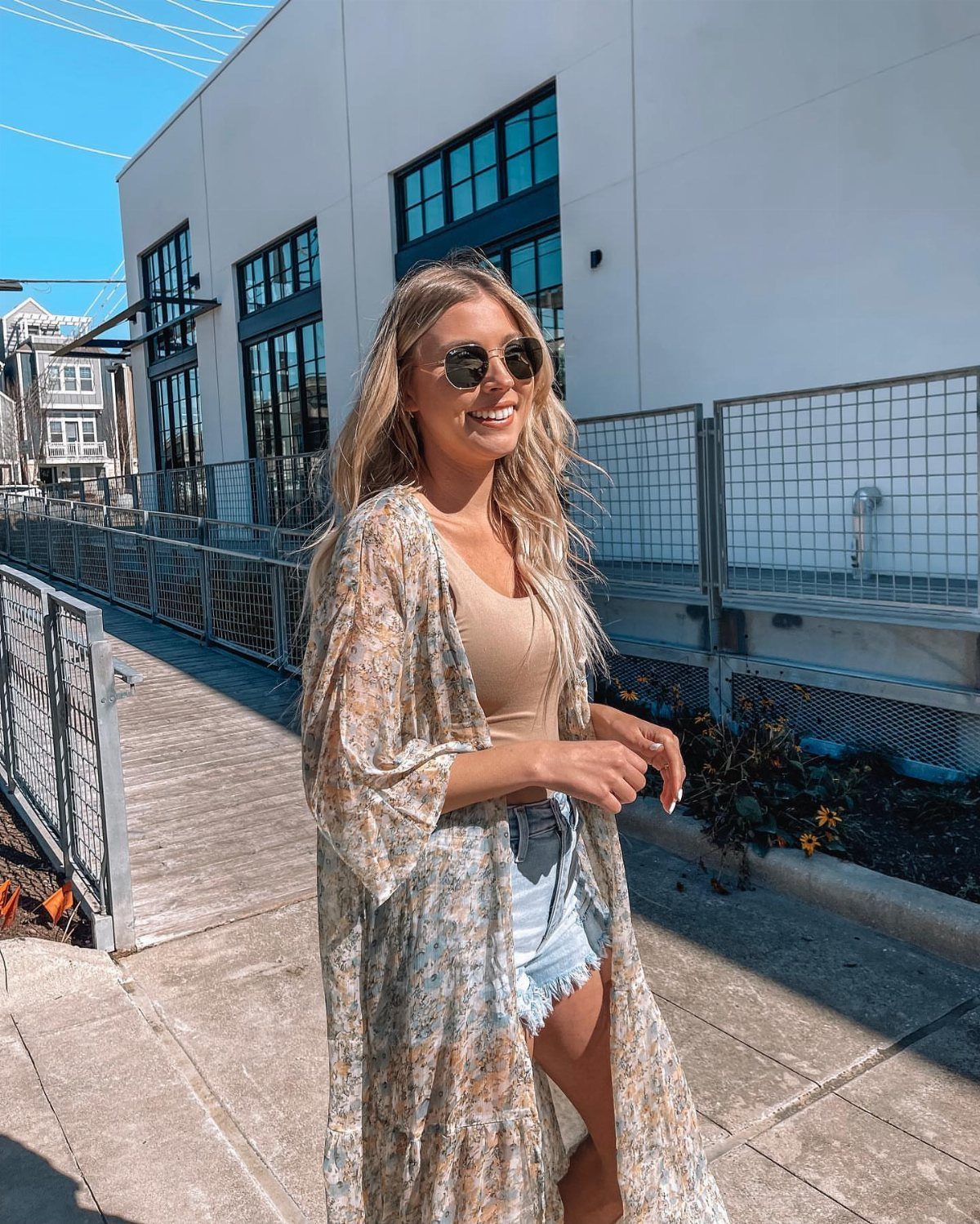 Woman wearing a floral kimono, tan top, and denim shorts outdoors in sunlight.