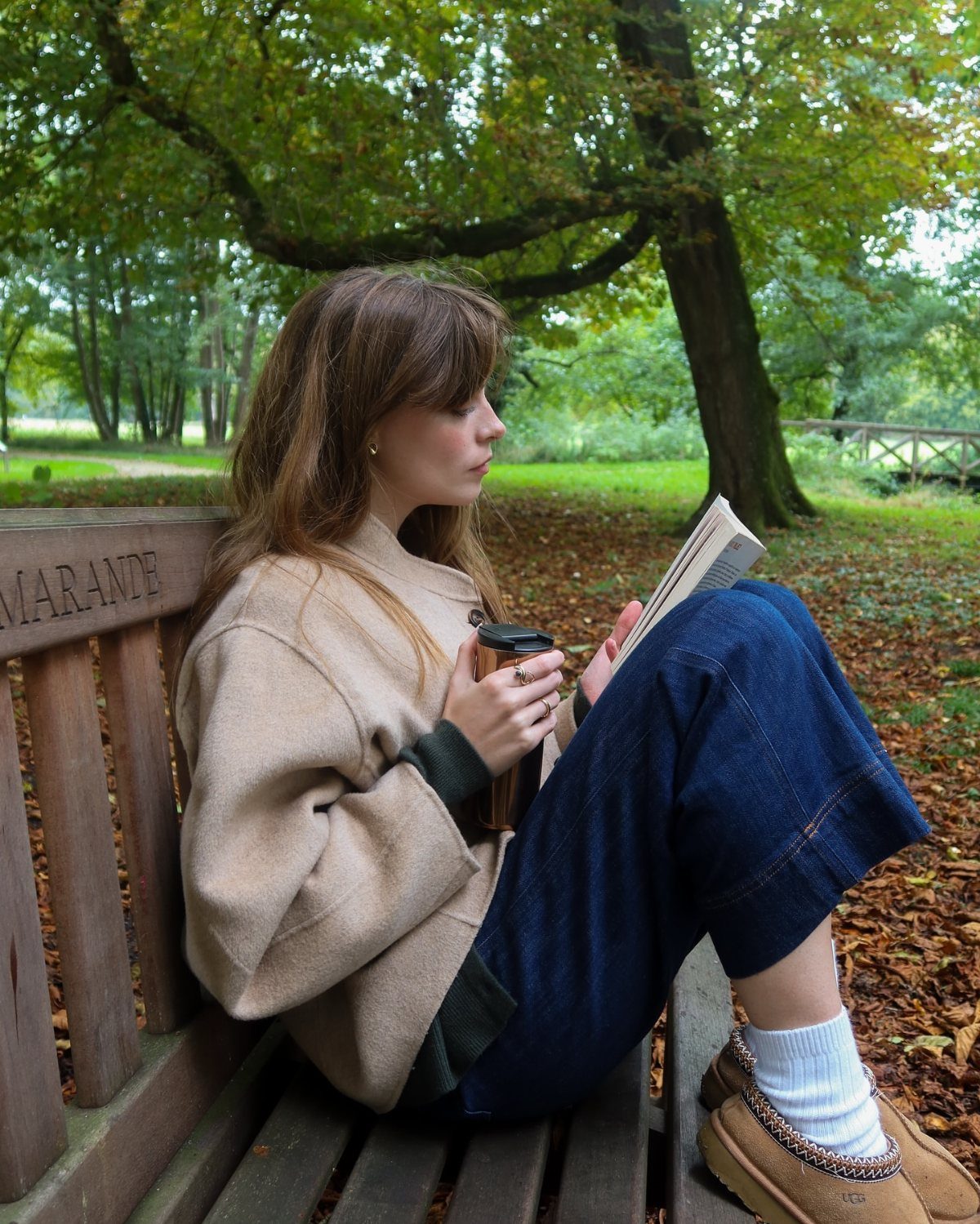 Woman in beige coat and wide-leg denim trousers reading a book on a bench.
