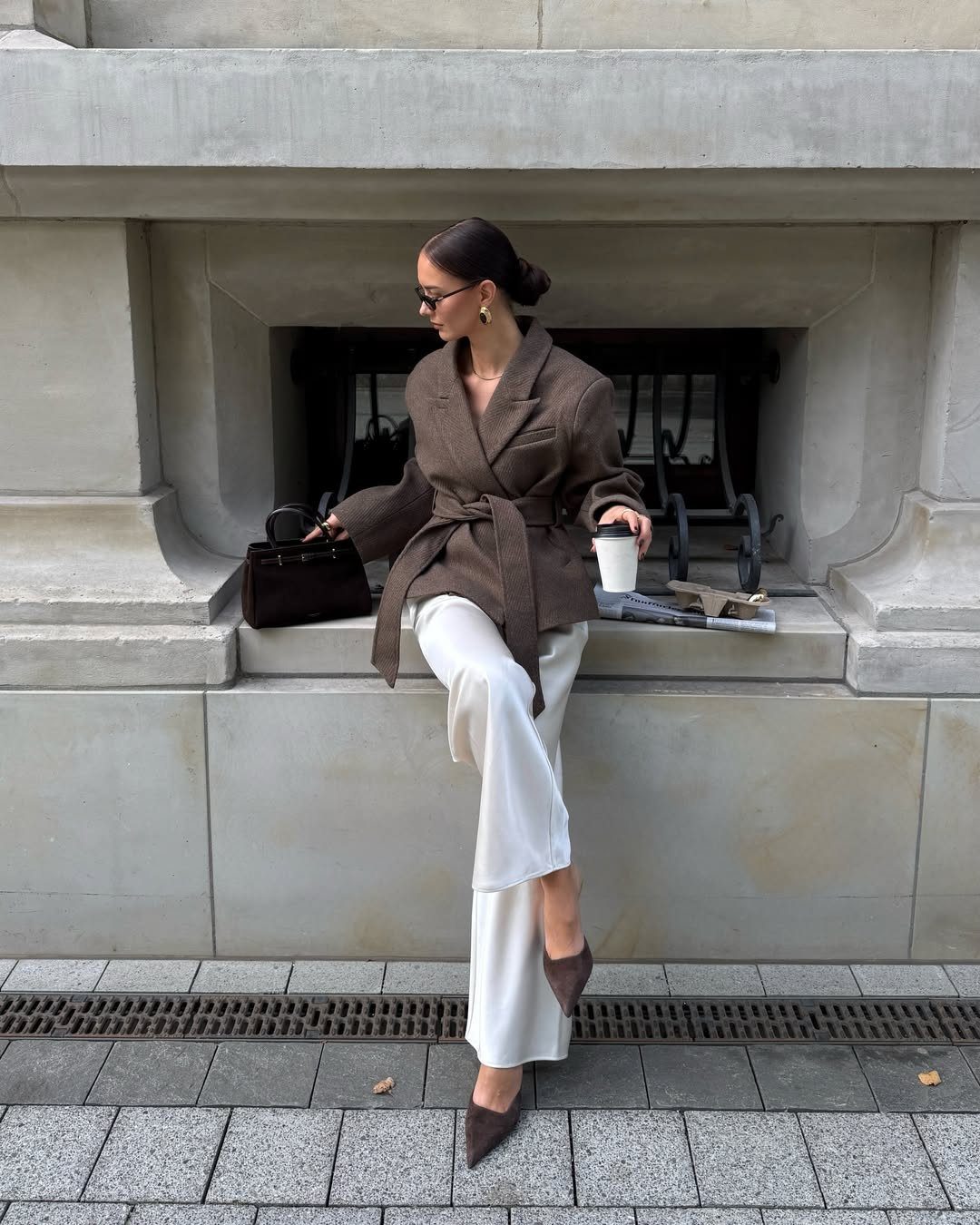Woman in brown belted blazer and white wide-leg trousers sitting on stone ledge.