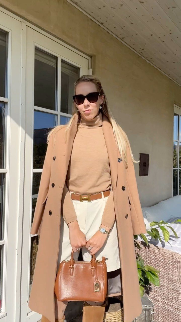 Woman in a camel coat and matching sweater with cream trousers and brown handbag.