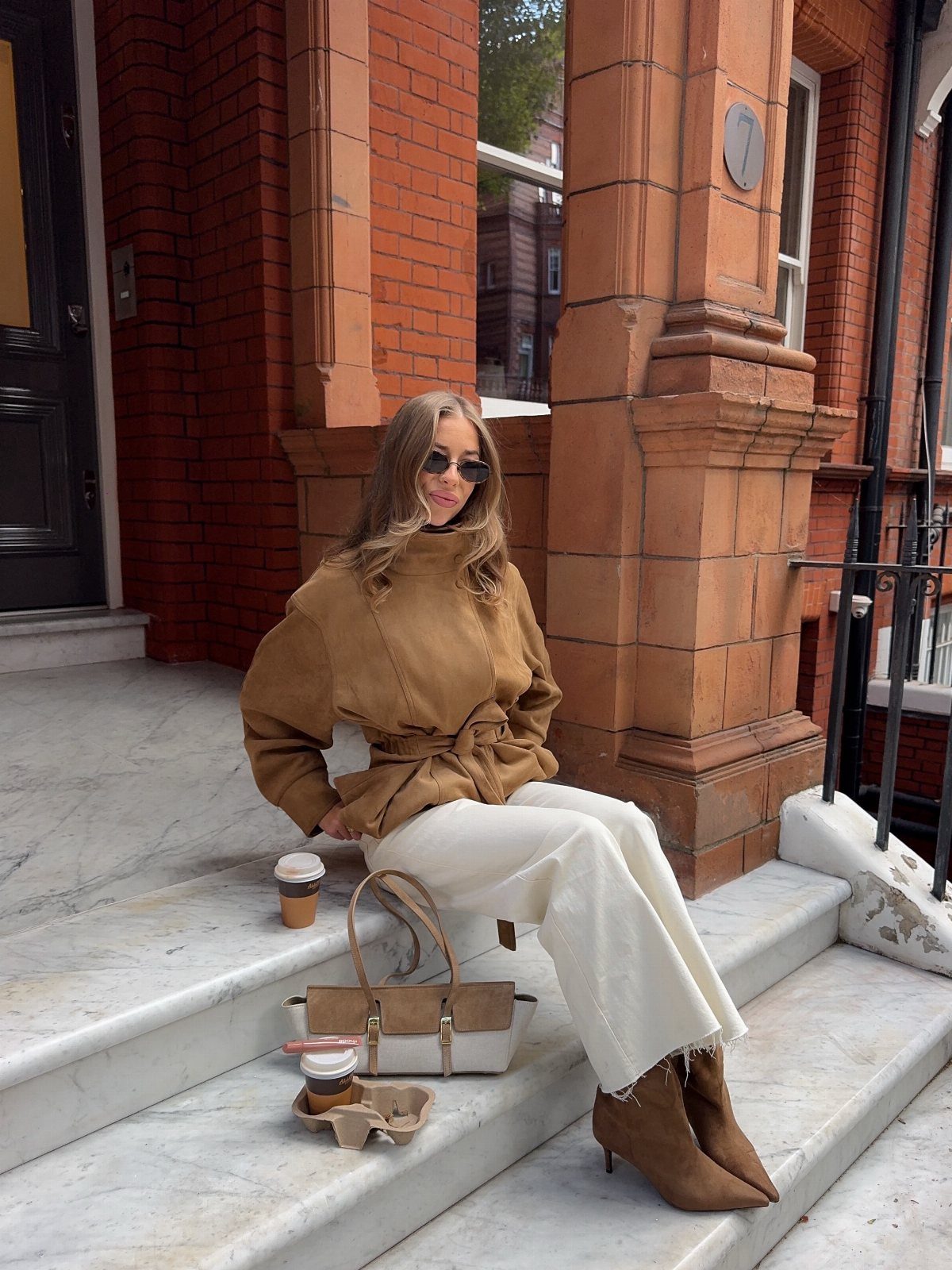 Woman in camel jacket and white wide-leg trousers sitting on marble steps.