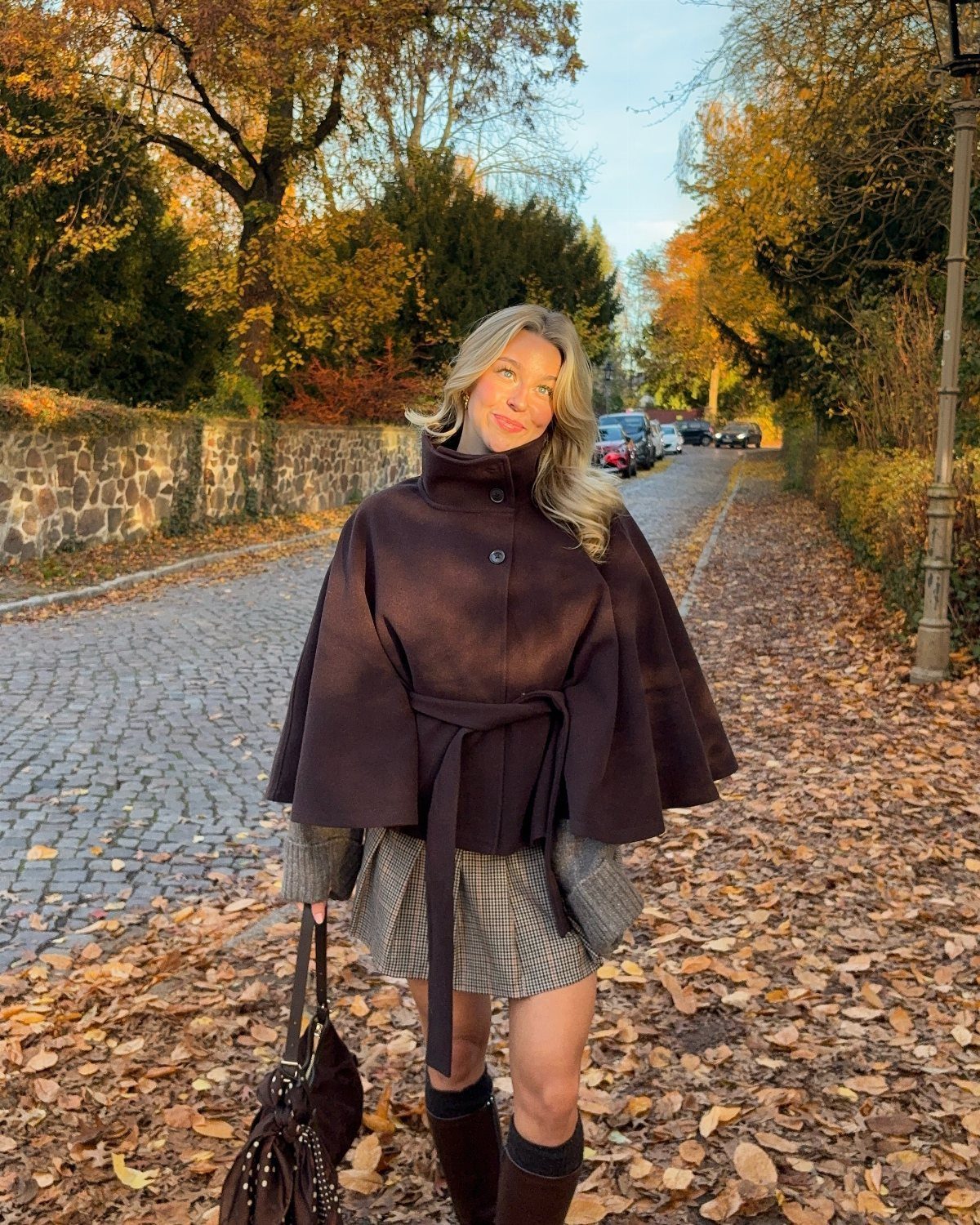 Woman wearing a brown belted cape and plaid skirt on a leaf-covered path.