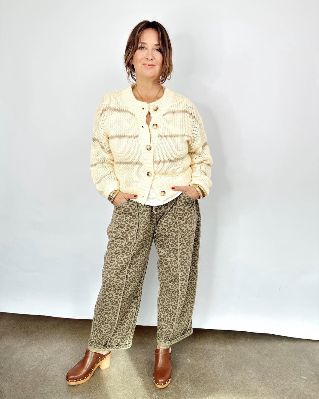 Woman wearing a cream cardigan and leopard print barrel-shaped trousers with brown clogs.