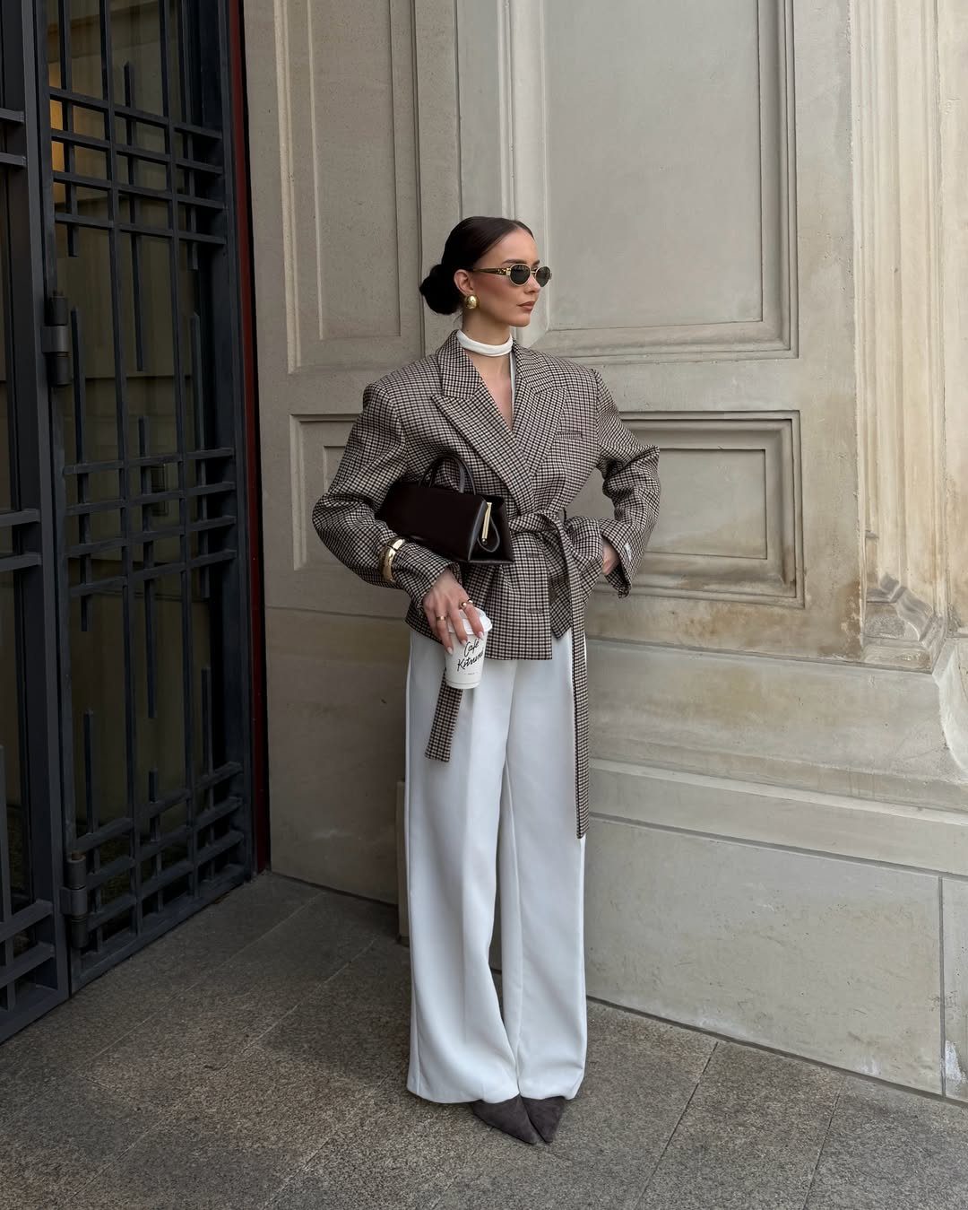 Woman wearing a checked blazer and wide-leg white trousers standing against a wall.
