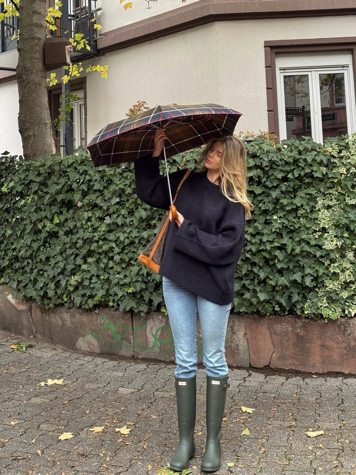 Woman in black sweater, blue jeans, and green rubber boots holding a plaid umbrella.