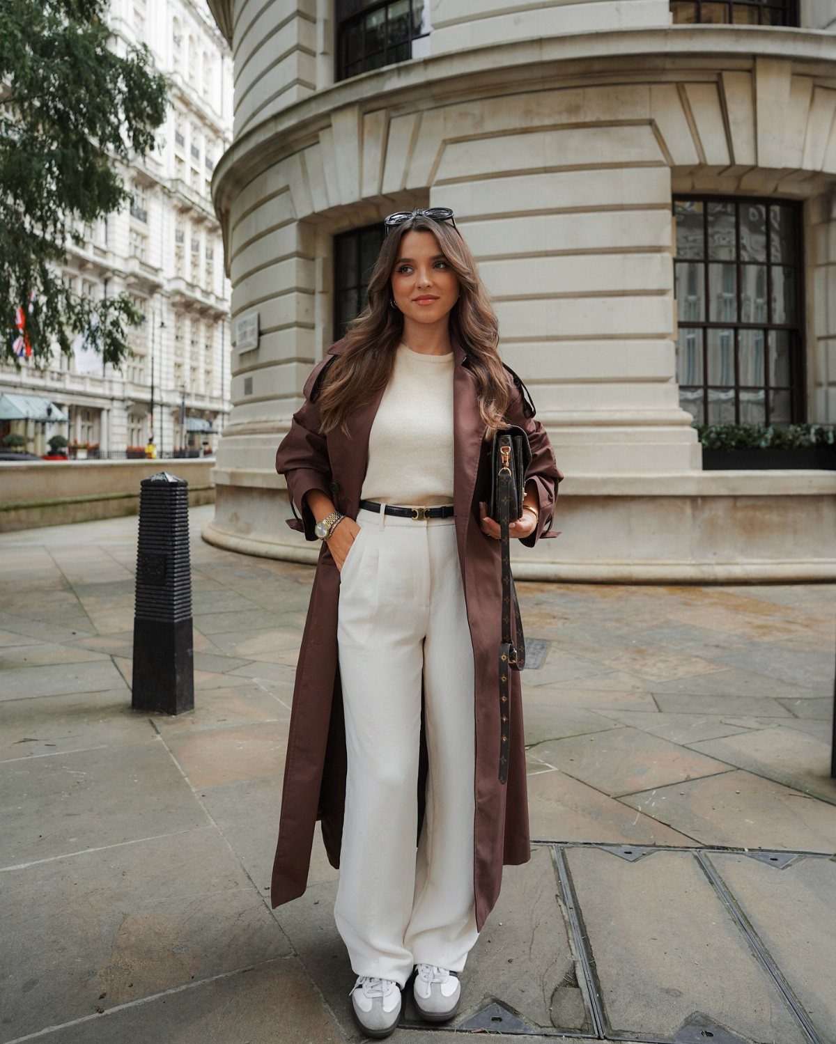 Woman wearing a long brown trench coat with cream trousers and white sneakers.