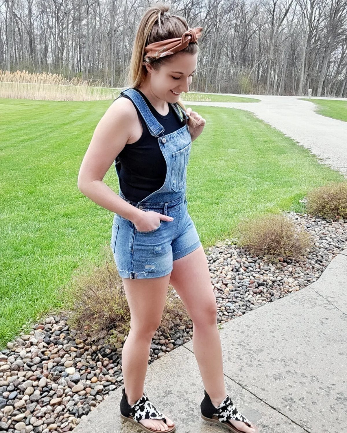 Woman wearing denim short overalls, black tank top, and cow-print slides on a path.
