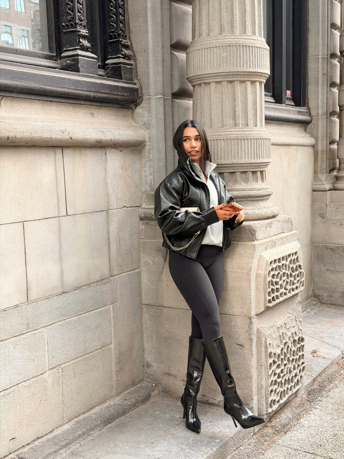 Woman in black leather jacket and leggings leaning against a stone building column.