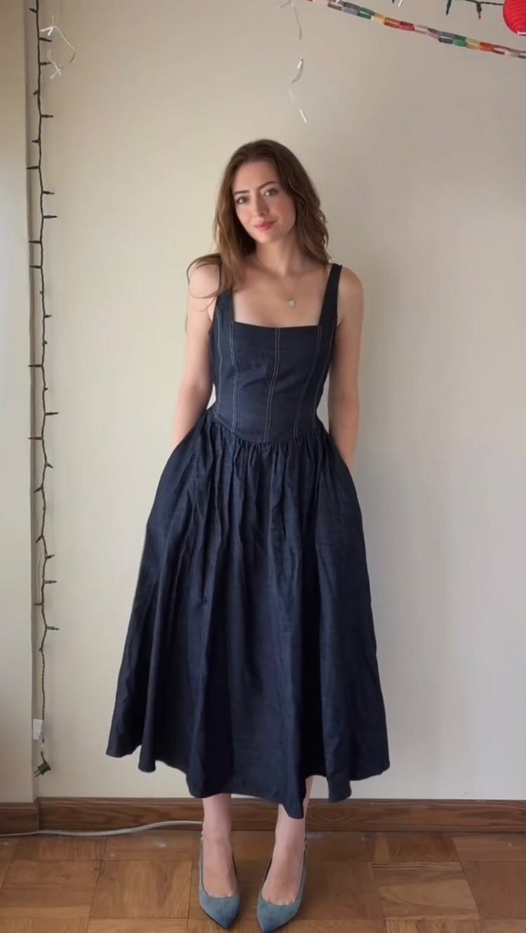 A woman posing in a dark blue sleeveless midi dress and light blue flats.