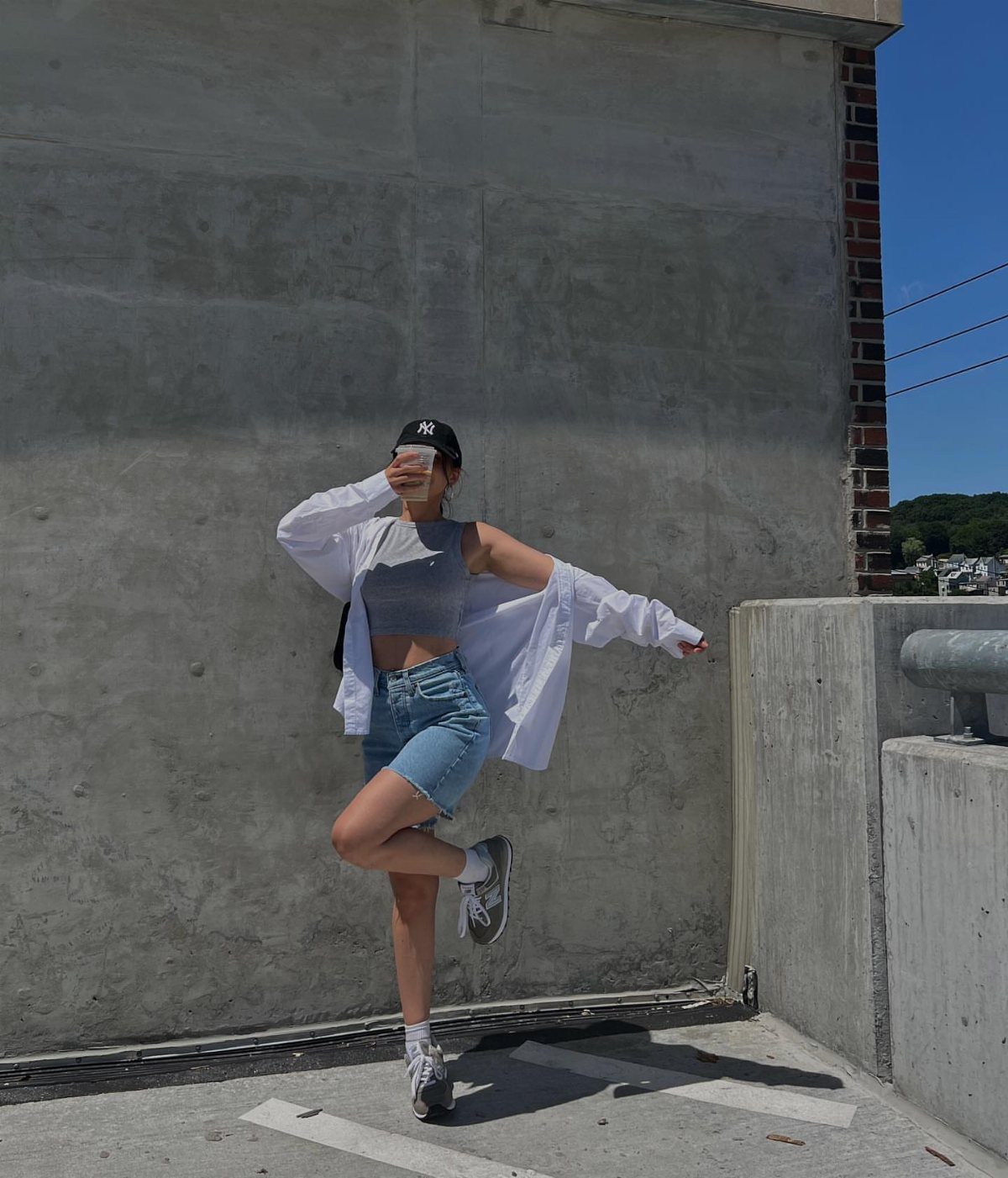 Person posing in denim shorts, grey crop top, and white shirt against a concrete wall.