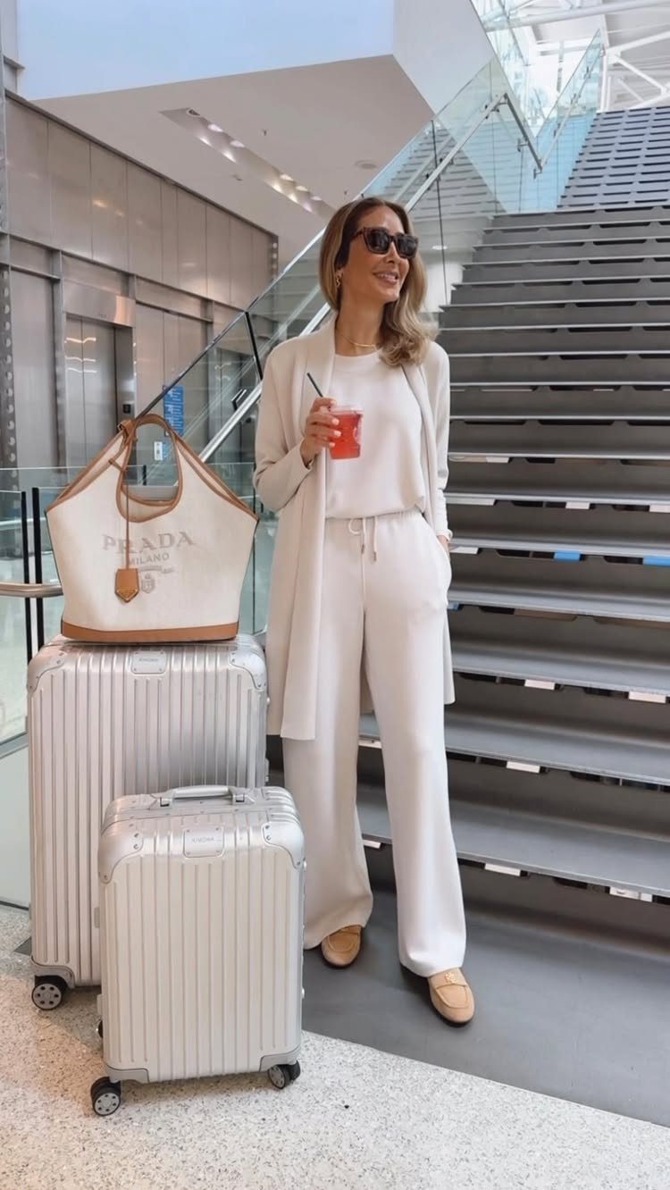 Cute Travel Outfits: 7 Ideas That Balance Style and Comfort 3 Woman in cream wide-leg lounge set standing with silver suitcases and a designer tote bag.