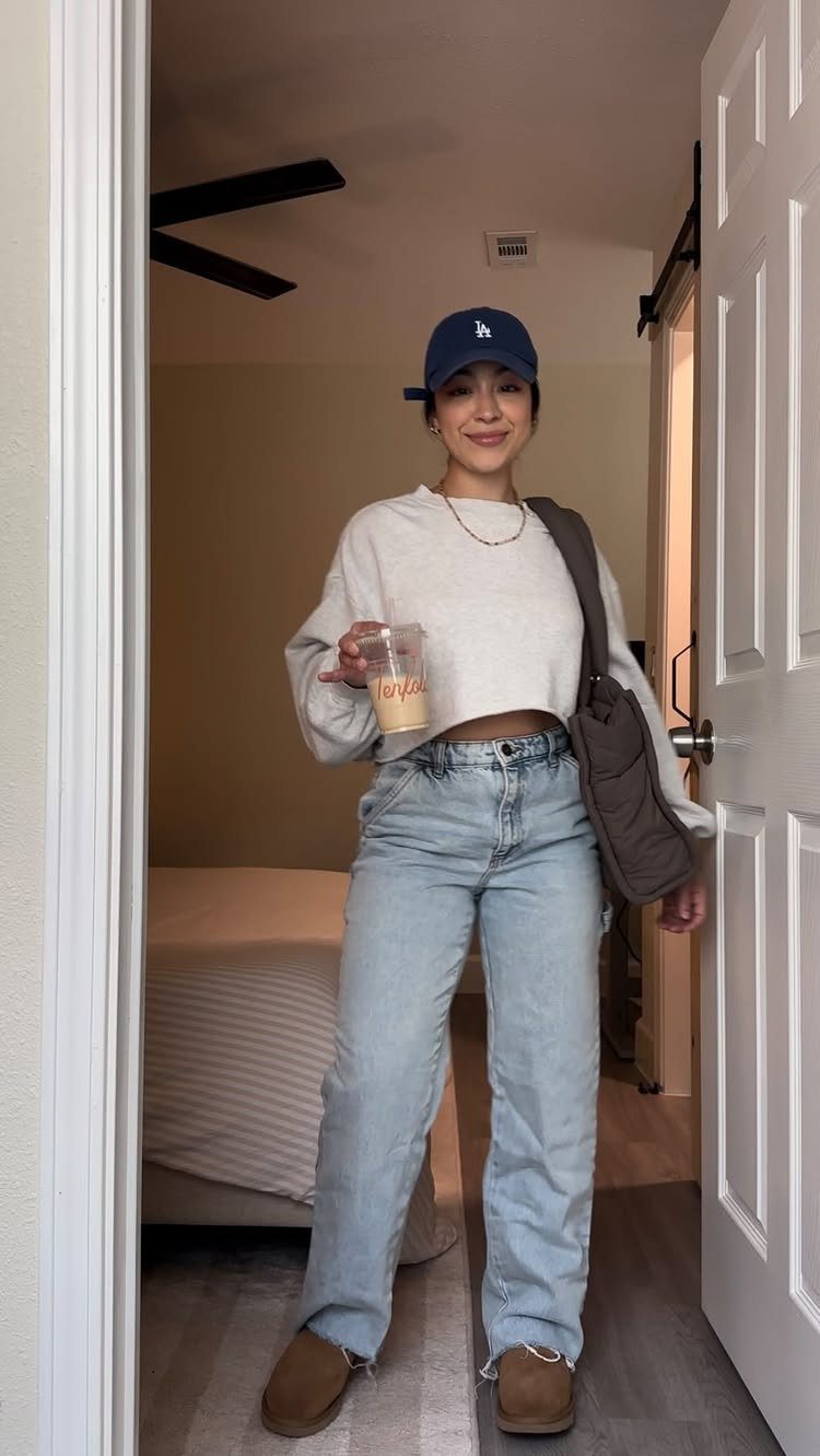 Smiling woman in a cropped sweater, light blue jeans, and a blue cap standing in a doorway.