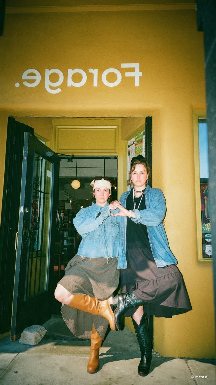 Two women posing in denim jackets, long skirts, and tall boots in front of a yellow storefront.