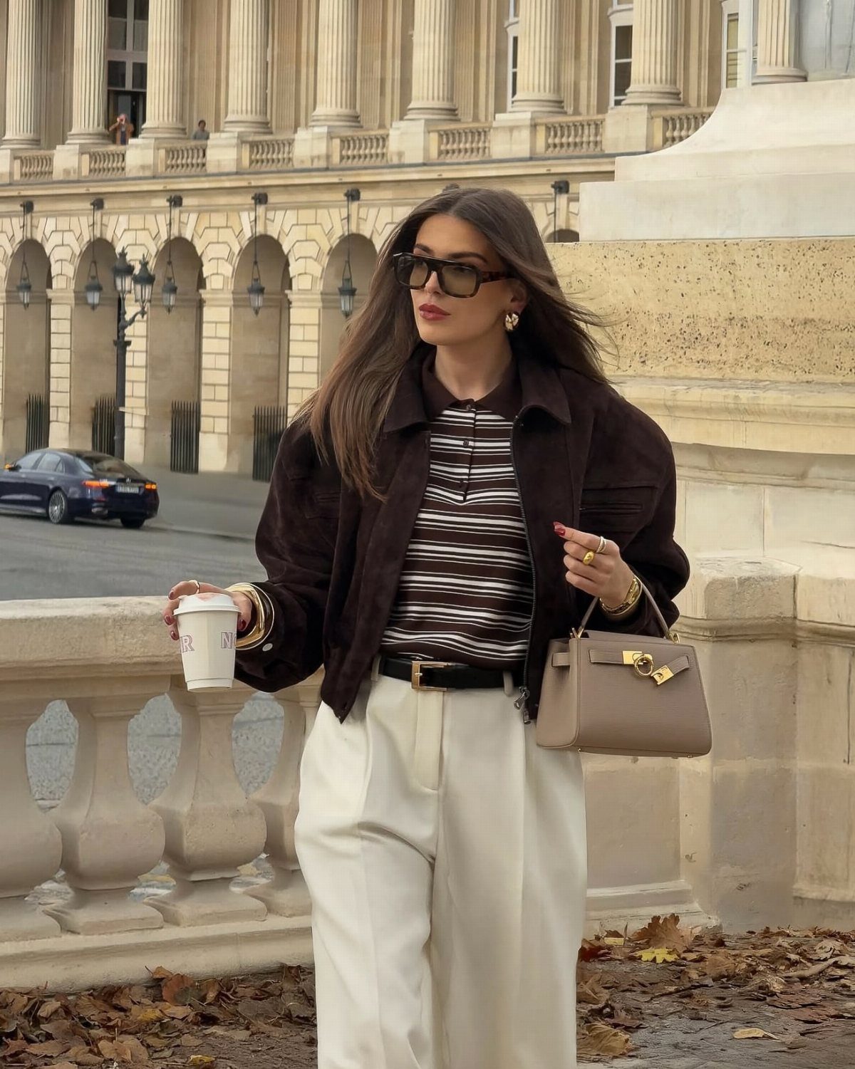 Woman in a brown suede jacket, striped shirt, and cream wide-leg trousers walking outdoors.