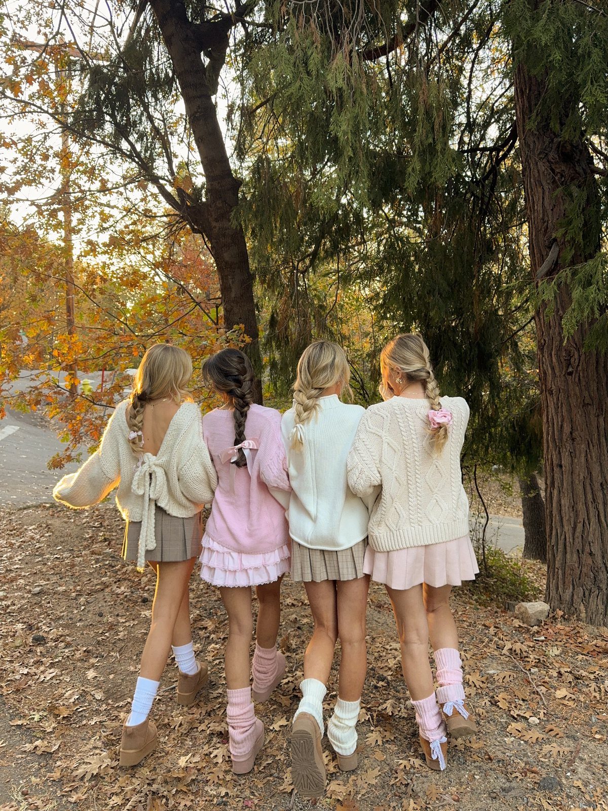 Four women in pastel sweaters and skirts standing among autumn trees and fallen leaves.