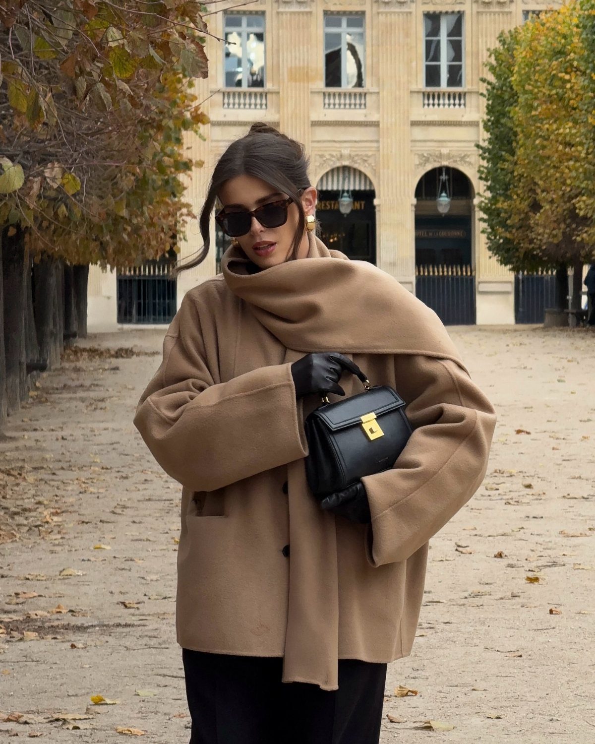 Woman wearing an oversized tan statement coat, black sunglasses, and black gloves while holding a small handbag.