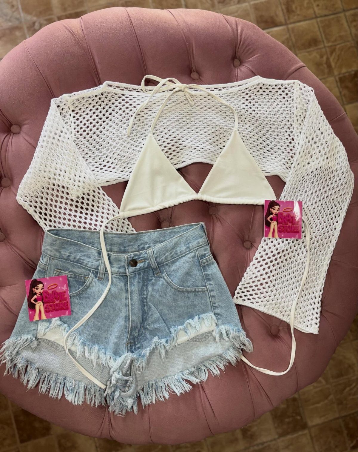 White crochet shirt, white bikini top, and distressed denim shorts on pink ottoman.