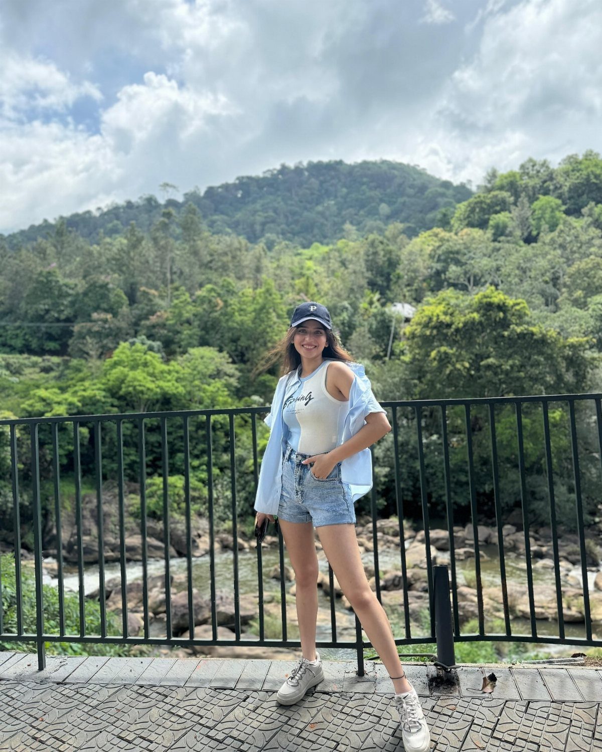 Woman in white tank top and denim shorts posing against a mountain backdrop