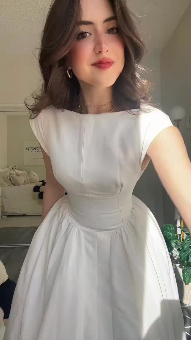 A young woman posing in a sleeveless white A-line dress with a boat neckline.