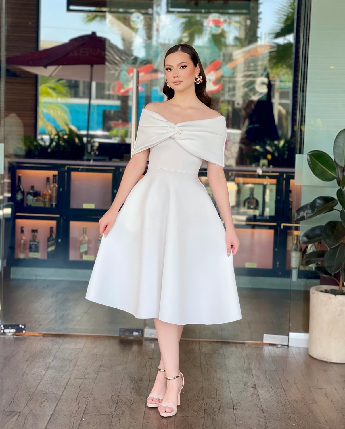 Woman wearing an elegant off-the-shoulder white satin midi dress standing on a wooden deck.