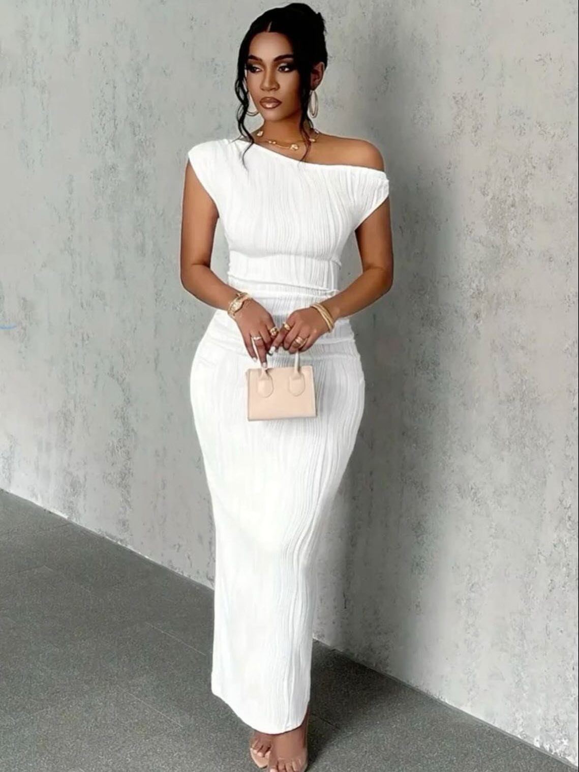 Woman wearing a white one-shoulder pleated maxi dress and holding a small beige handbag.