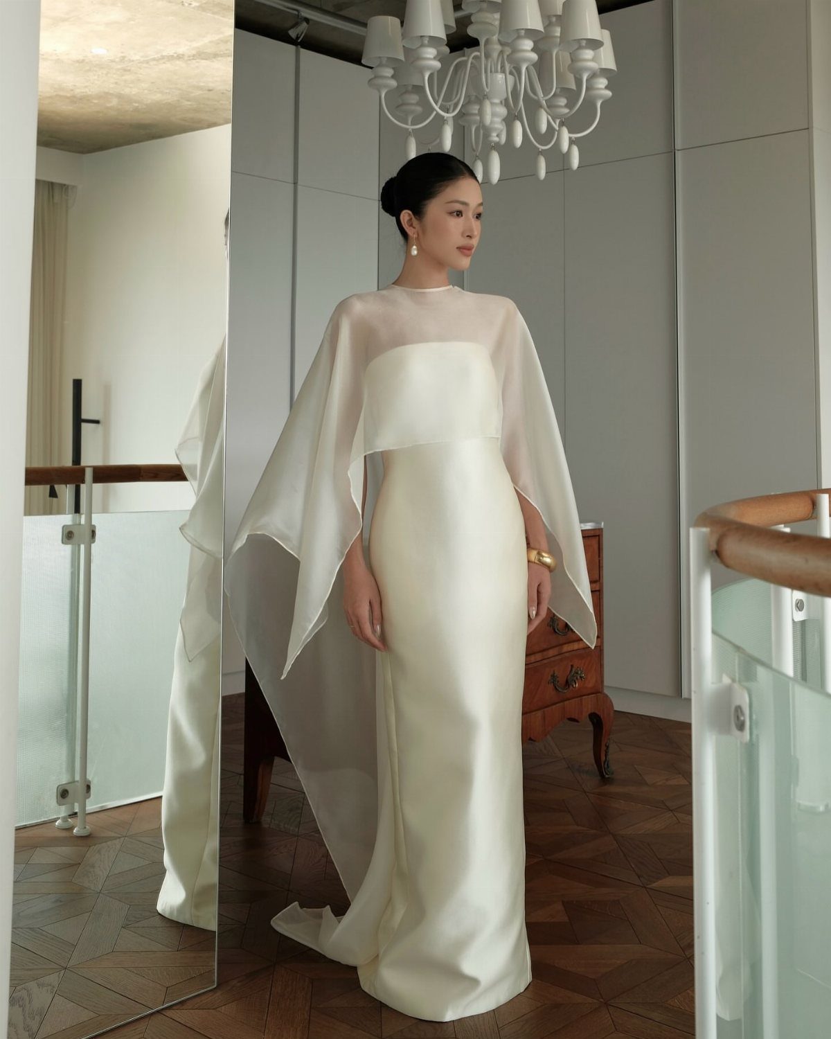 A woman in a long white satin gown with a sheer flowing cape stands in a room.