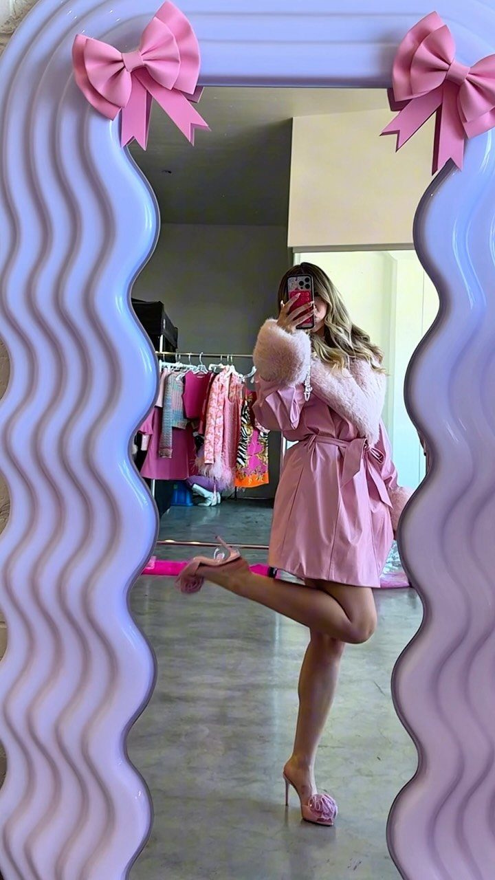 Woman in a pink fur-trimmed robe taking a mirror selfie in a room with clothing racks.