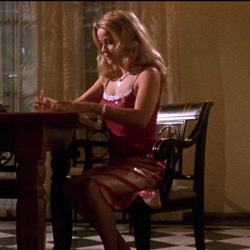 Blonde woman in a pink tank top and patterned skirt sitting at a wooden table.