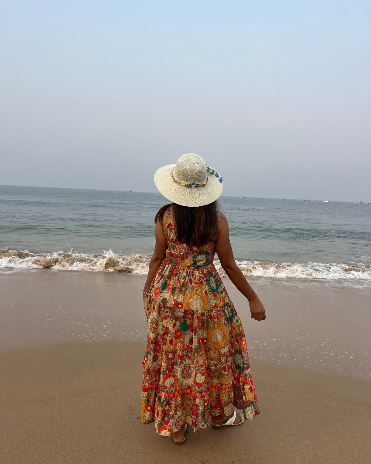 Fall Beach Outfits: 20 Ideas for Your Coastal Getaway 20 Woman in a colorful floral maxi dress and sun hat standing on a beach.