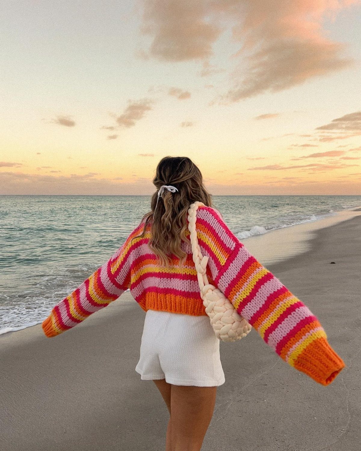 Fall Beach Outfits: 20 Ideas for Your Coastal Getaway 10 Woman in a colorful striped sweater and white shorts standing on a beach.