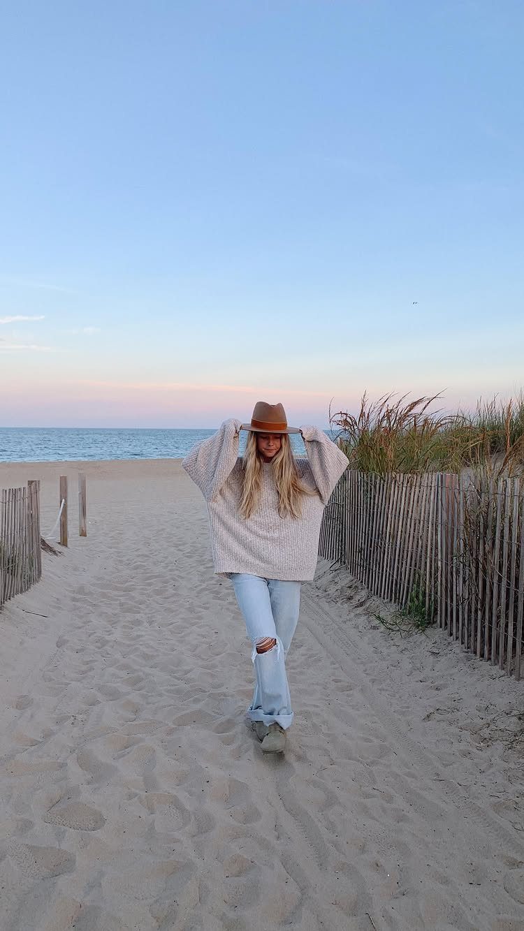 Fall Beach Outfits: 20 Ideas for Your Coastal Getaway 19 Woman wearing a cream knit sweater, brown hat, and jeans on a sandy beach.