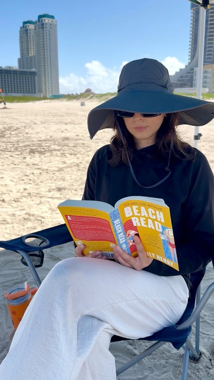 Fall Beach Outfits: 20 Ideas for Your Coastal Getaway 13 Woman reading a book on a beach wearing a wide-brimmed hat and sunglasses.