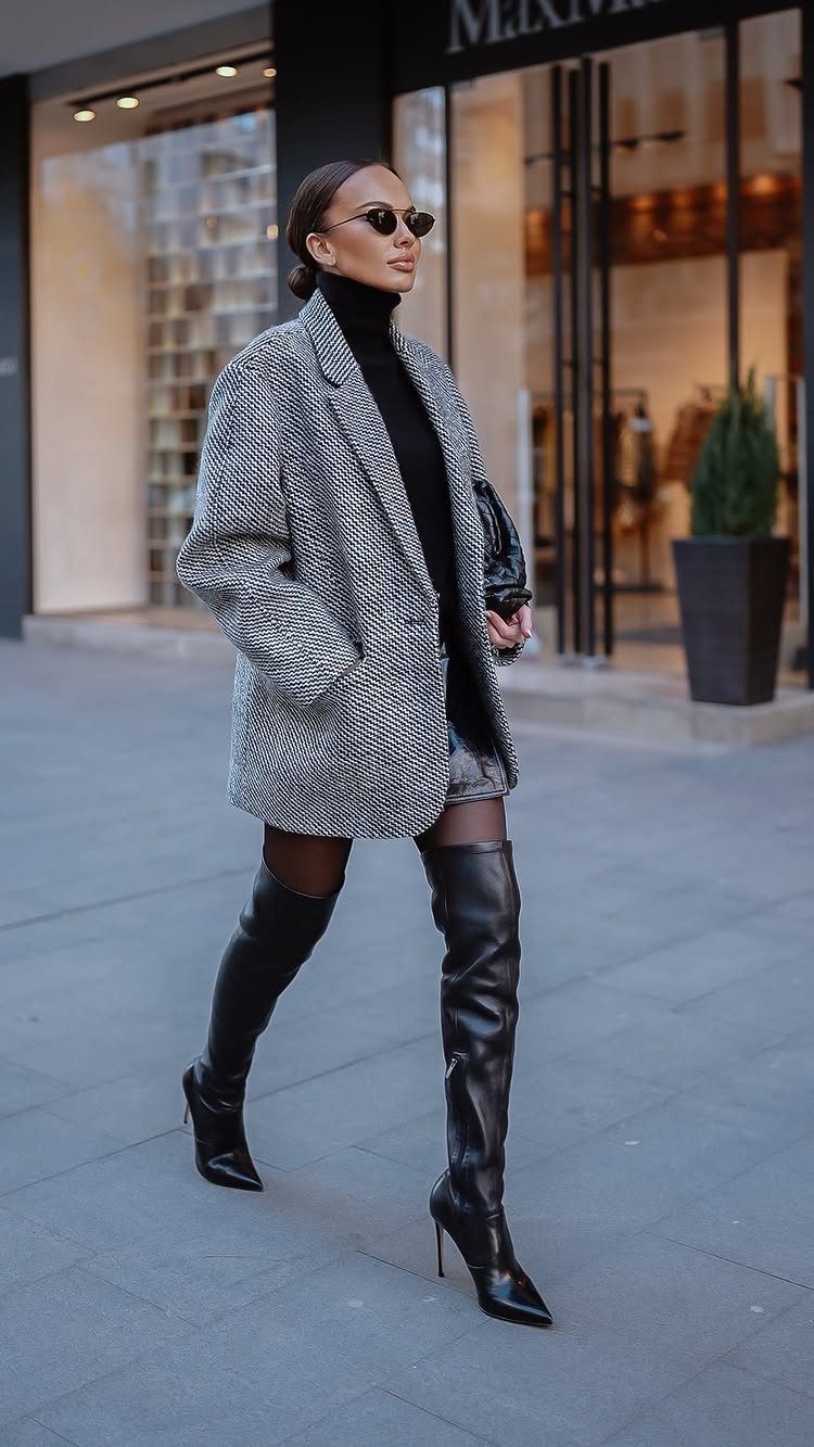 Woman in oversized grey blazer and black leather knee-high boots on city street.