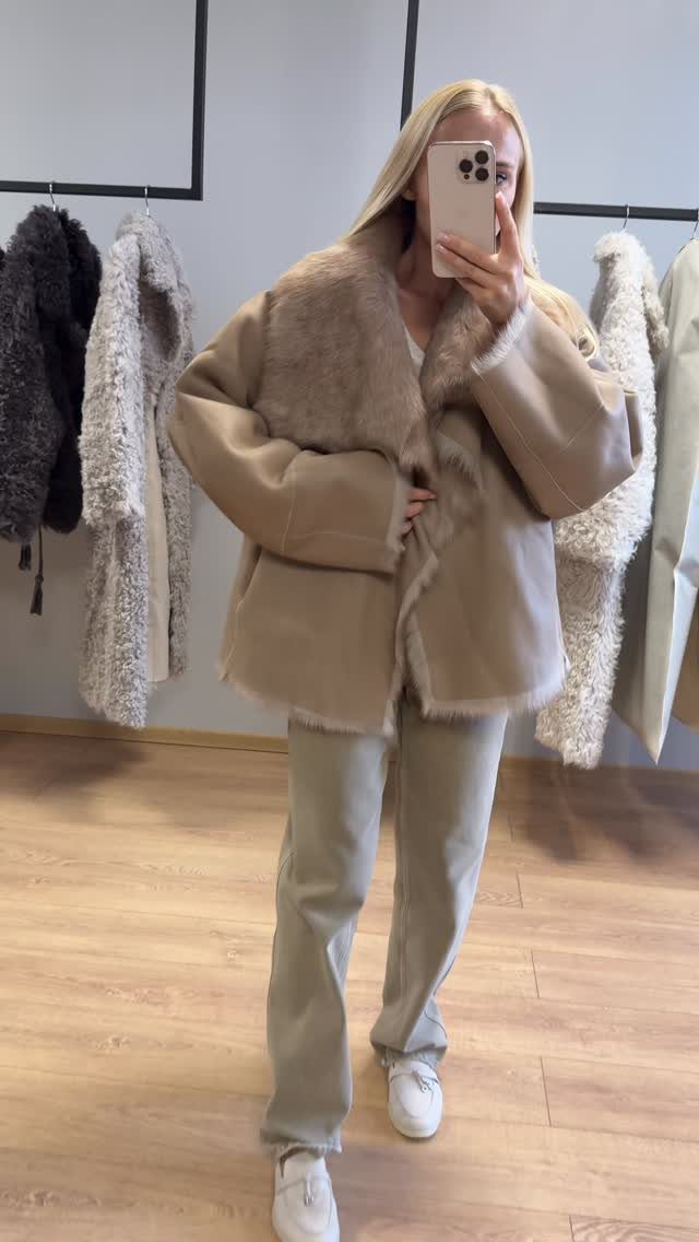 Woman in a beige faux-fur trimmed jacket and olive trousers taking a mirror selfie.
