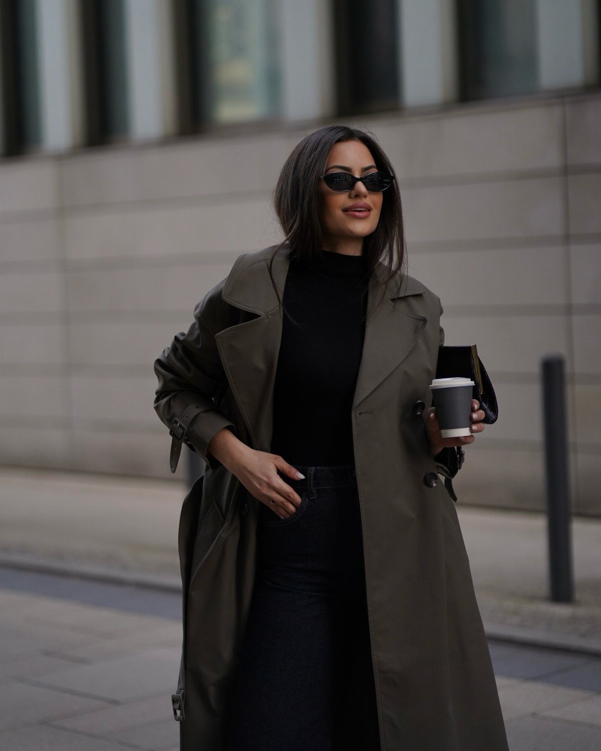 Woman in olive green long coat and dark-wash denim jeans holding coffee.