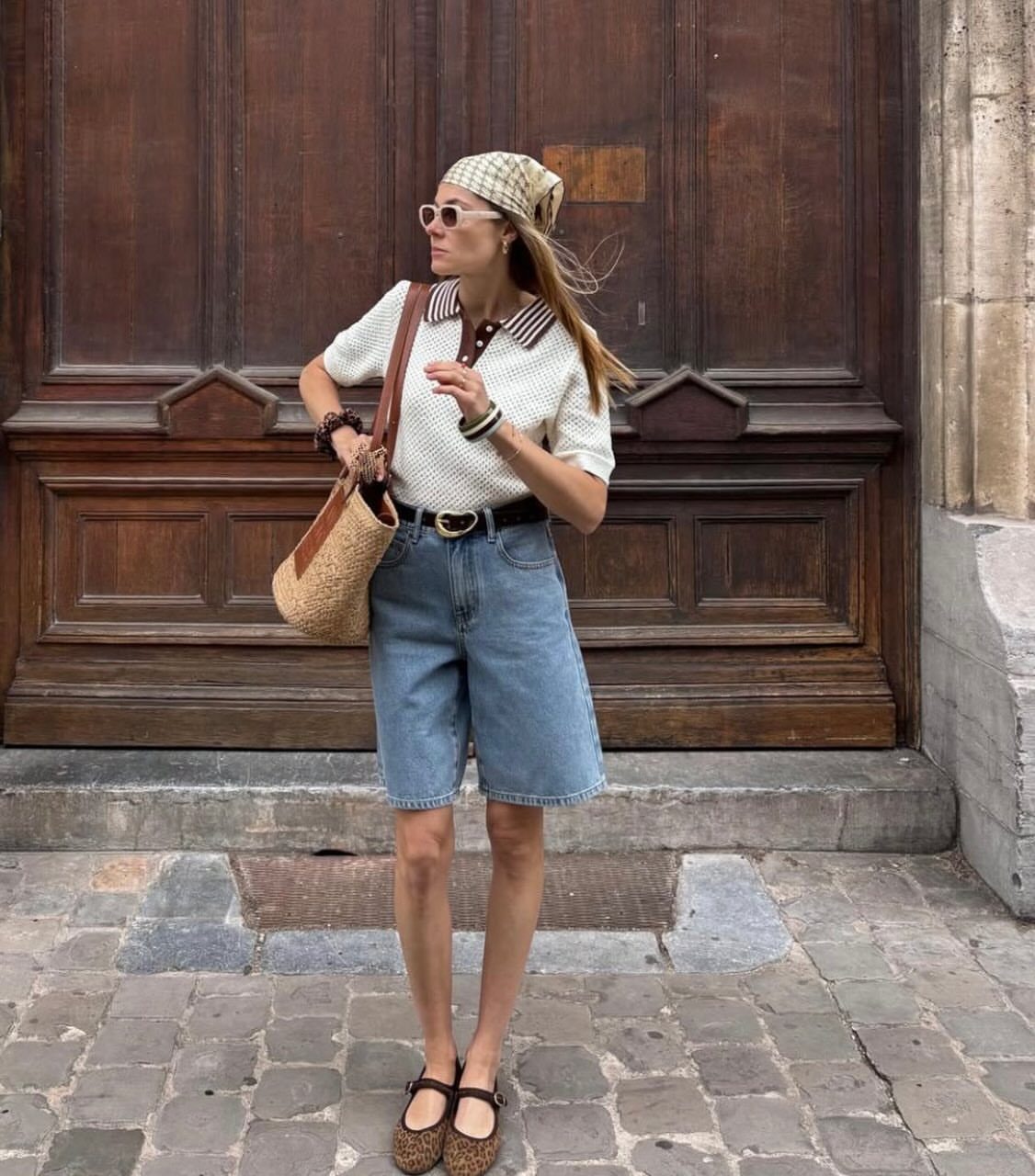 Woman wearing long light-wash denim shorts and a white knit top standing outdoors.