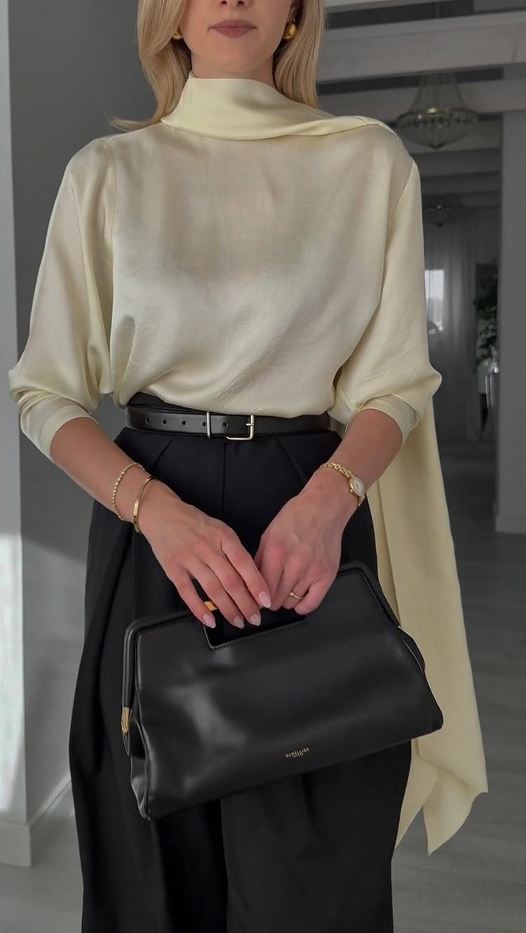 Woman in cream satin blouse and black trousers holding a black leather handbag.