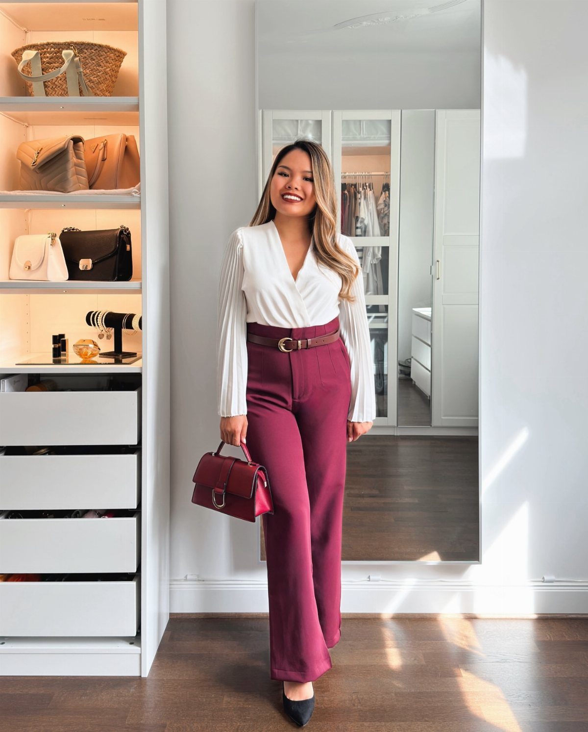 Woman wearing a white blouse and burgundy wide-leg trousers in a bright room.