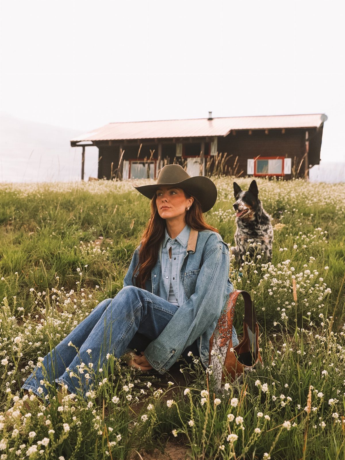 Woman in denim jacket and cowboy hat sitting in a wildflower field with dog.