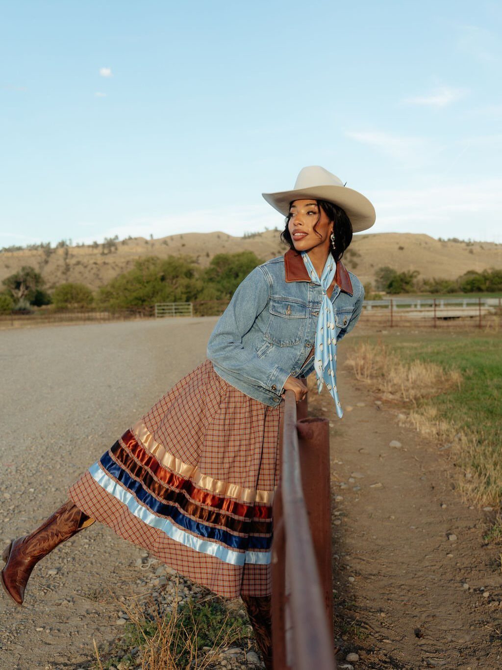 Woman in a short denim jacket, plaid skirt, and cowboy hat posing outdoors.