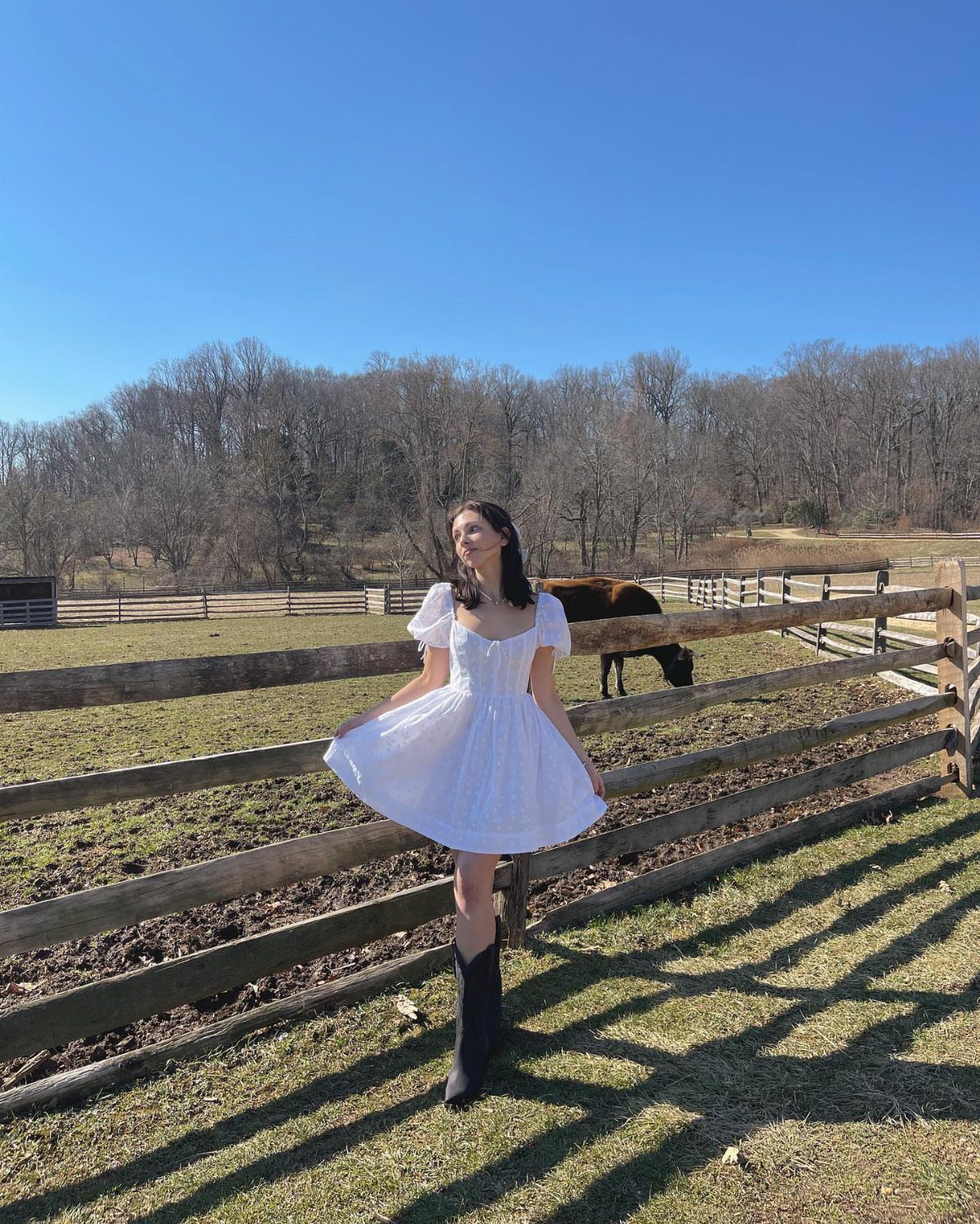 Woman in a white sweetheart dress and black boots standing by a farm fence.