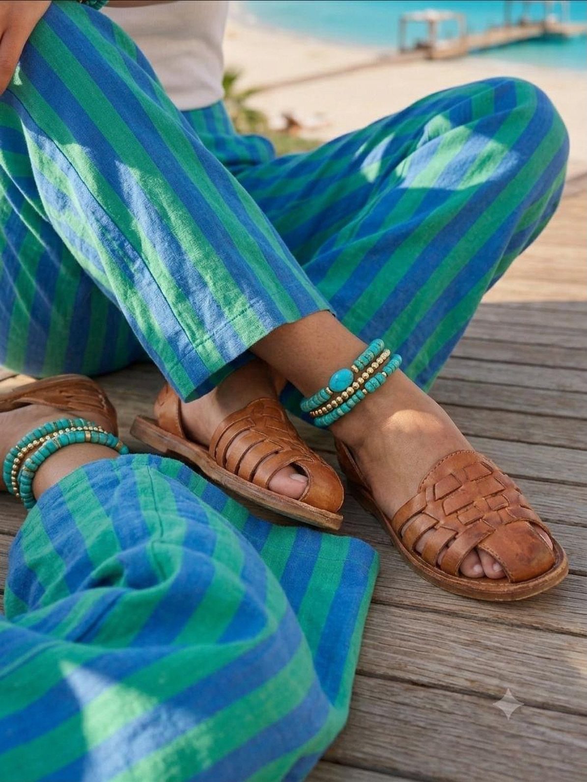 Blue and green striped beach pants with brown woven leather sandals and beaded anklets.