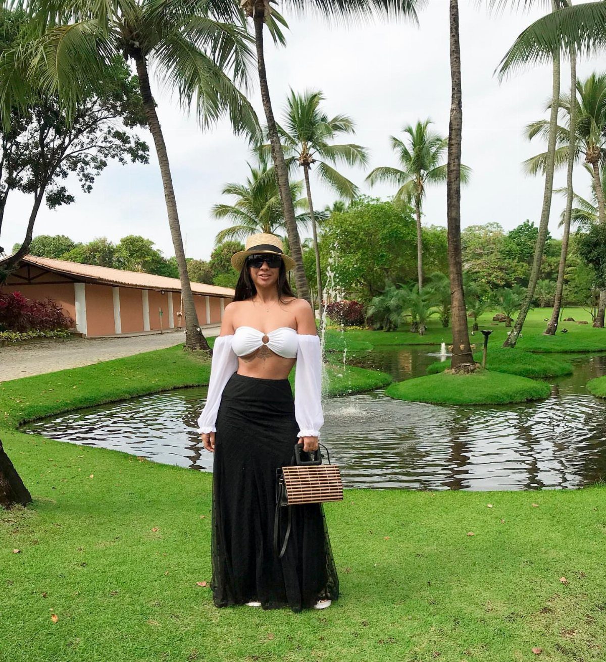 Woman in white crop top and black maxi skirt standing by a tropical pond.