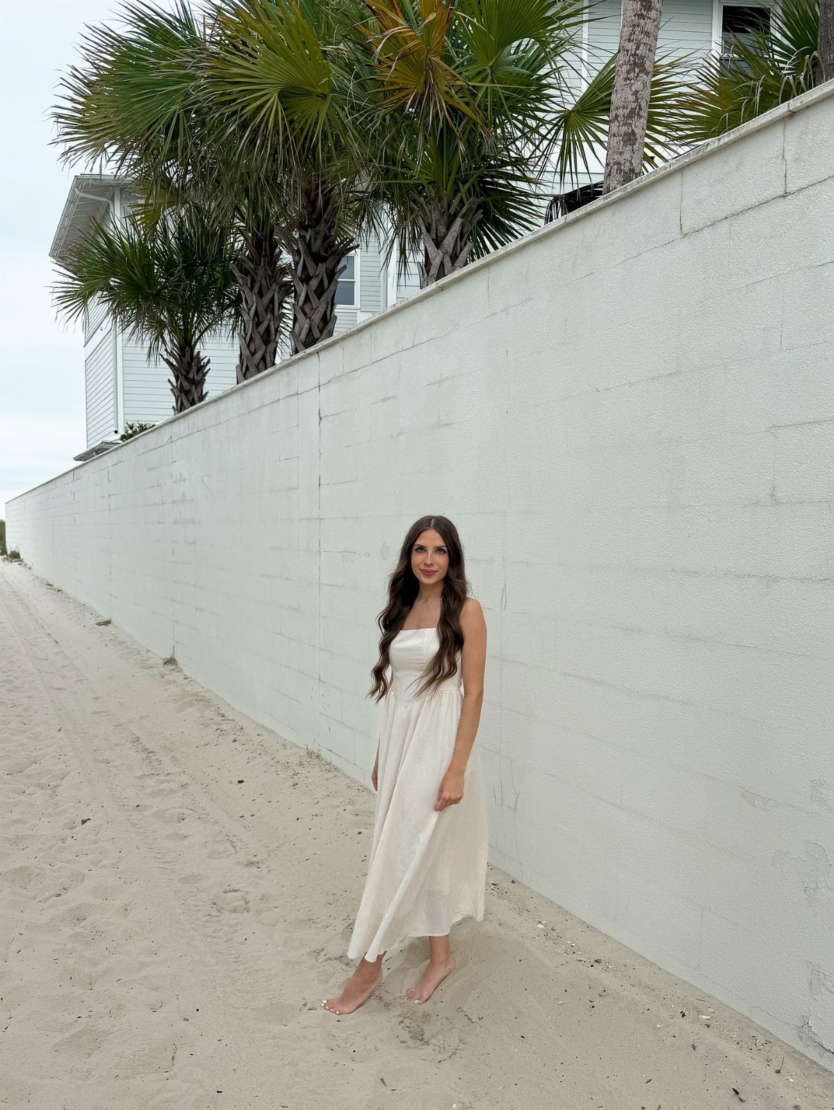 Woman in a long white sleeveless sundress standing on a sandy beach