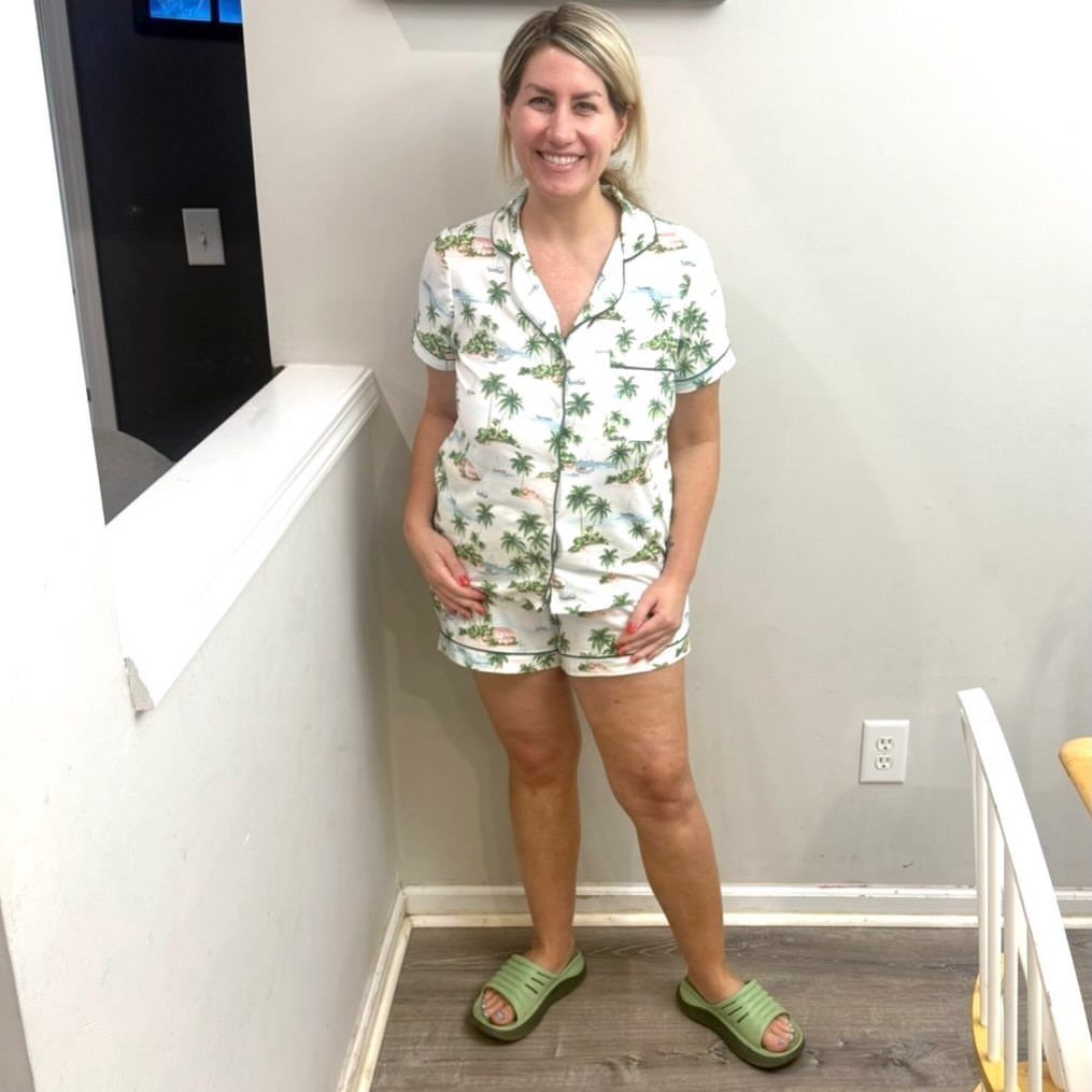 Woman wearing white tropical print pajama set and green slides in a home.